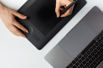 Top view photo of a female hand drawing on a graphic design tablet, stylus and a laptop on a white...