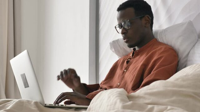 Concentrated African-American man with glasses in pajama works on laptop recovering after covid on comfortable bed in room closeup