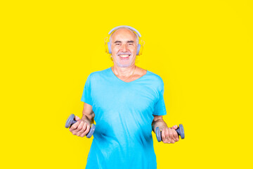 Elderly man training using dumbbells on yellow background listening music headphones