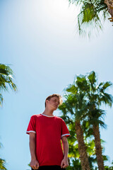 cute young teenage boy posing and having fun on the beach under palm trees in red T-shirt . Trendy boy posing. Funny and positive man in sunglasses