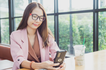 Cheerful Asian businesswoman in pink suit with eyeglasses sitting and holding smartphone at wooden table inside modern restaurant cafe. Beautiful cute young smiling glasses girl, using mobile phone