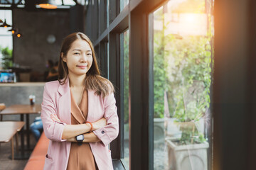 Young Asian businesswoman in pink suit standing with arms folded in coffee shop, confident  herself, pride owner of a business modern restaurant. Smiling beautiful cute girl with happy face in cafe