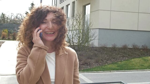 Curly-haired middle-aged woman walks along a deserted city street. She speaks on the phone with the one who is waiting for her at the appointed place. Woman waves her hand in greeting.