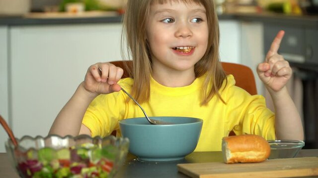 Close up of little smiling girl sits by table and eats tomato soup with croutons and cheese. Child looks at camera and has lunch on kitchen background.