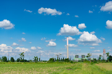 Fototapeta na wymiar Thermoelectric power plant on the horizon, in a rural environment, with green cultivated field used in agriculture. Blue sky with white clouds on the background, Italy.
