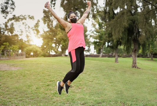 Happy curvy woman wearing safety face mask and jumping outdoor at city park while listening music playlist with wireless headphones