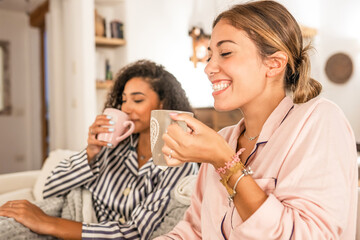Fototapeta na wymiar Couple of cohabiting girls in pajamas relaxing under blanket on cozy sofa drink a cup of tea while having fun talking and joking with each other. Real life moments of diverse multiracial gay couple