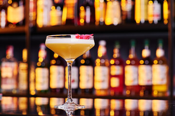 Elegant female yellow cocktail with white foam, decorated with pink flower petal on dark table against the background of blurred bar with expensive alcohol. Close-up. Space for inscriptions