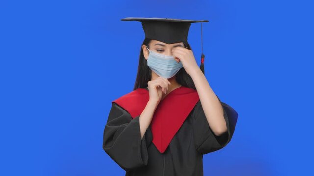 Portrait of female student in cap and gown graduation costume puts medical protective mask. Young brunette woman posing in studio with blue screen background. Close up. Slow motion ready 59.94fps.