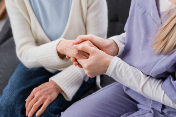 Fototapeta premium partial view of young social worker holding hand of elderly woman while sitting at home