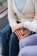 partial view of nurse sitting near senior woman and touching her hand, blurred background