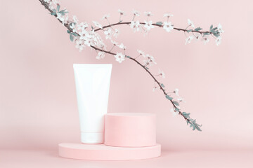 Blank white cosmetics tube on round platform podium stand and spring flowering tree branch with white flowers on pastel background. Front view