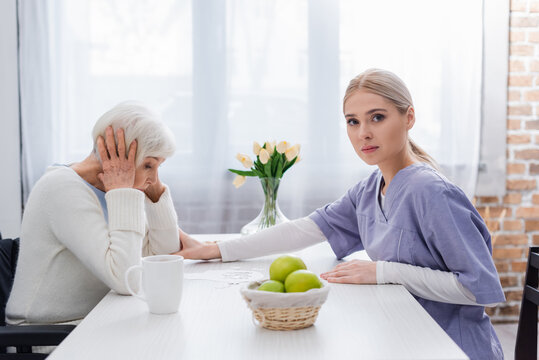 young nurse looking at camera while calming upset senior woman near jigsaw puzzle