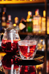Beautiful transparent teapot and cup of red fruit tea on a shiny dark table against the background of a blurred bar. Close-up. Space