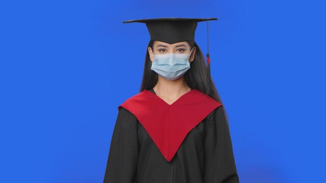 Portrait of female student in cap, gown graduation costume and medical mask looking at camera. Young brunette woman posing in studio with blue screen background. Close up. Slow motion ready 59.94fps.