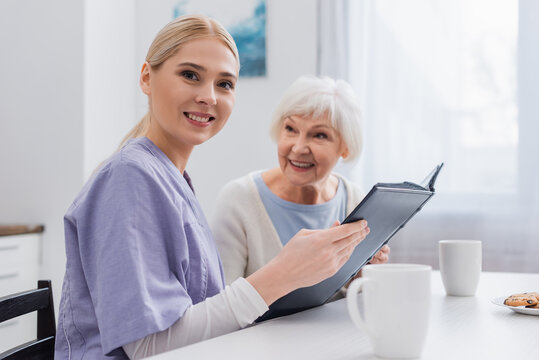 happy social worker looking at camera while holding photo album near aged woman