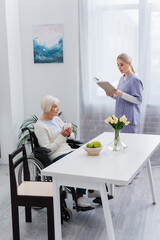 young nurse in uniform reading book to senior disabled woman sitting in kitchen with cup of tea