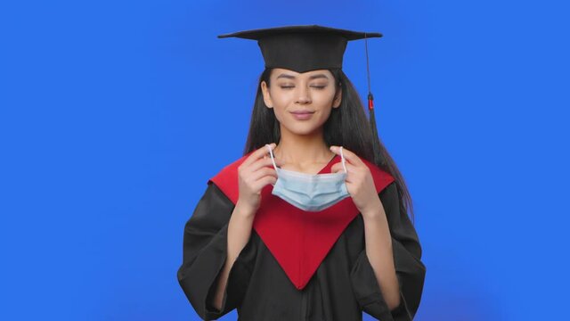 Portrait of female student in cap and gown graduation costume removes protective medical mask. Young brunette woman posing in studio with blue screen background. Close up. Slow motion ready 59.94fps.