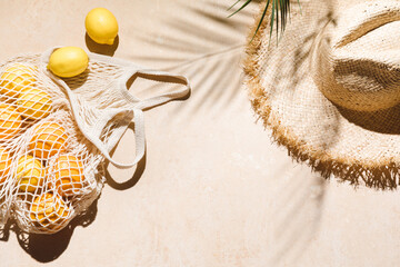 Summer flat lay on beige background. Straw hat and lemon fruits in eco friendly mesh shopping bag. Trendy palm shadow and sunlight, sun. Minimal summer travel fashion concept - obrazy, fototapety, plakaty