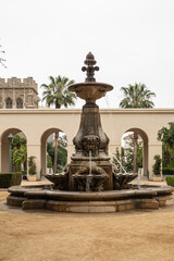 Obraz premium Pasadena, CA, USA - May 11, 2021: Bronze fountain on sandy courtyard and green foliage garden of historic City Hall under silver sky. Beige stone constructions in back.