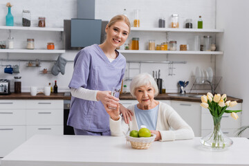 Obraz na płótnie Canvas young nurse and senior woman smiling at camera while holding glass of water and medication in kitchen