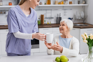 young social worker giving cup of tea to happy elderly woman in kitchen