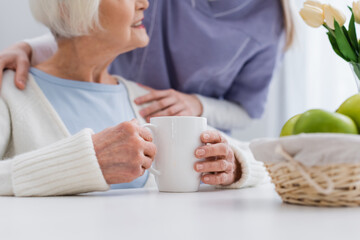 Obraz na płótnie Canvas cropped view of nurse embracing shoulder of smiling senior woman near cup of tea
