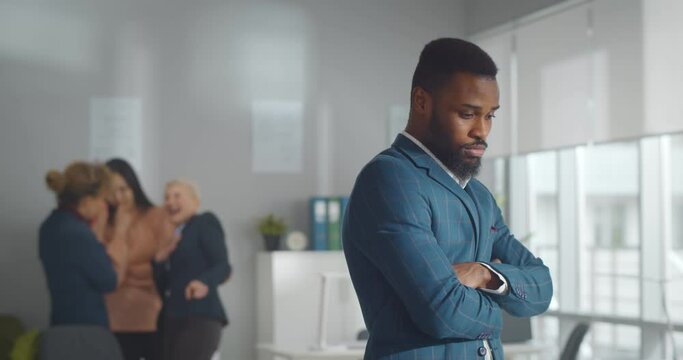 Portrait of sad african businessman standing in office with female colleagues laughing at him on background