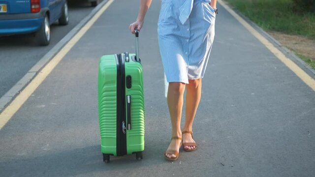 Closeup of young woman walking on city street sidewalk with green suitcase on summer day. Travelling and vacation concept.