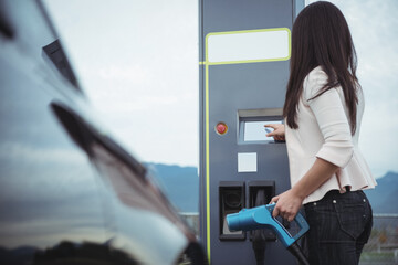 Caucasian woman holding charging cable for electric car keying in code in charging station