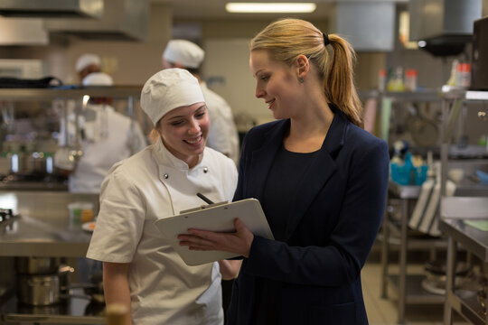 Caucasian female restaurant manager and chef using tablet and smiling in professional kitchen