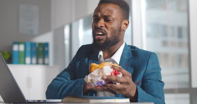 African businessman sitting at workplace eating burger and having heart attack