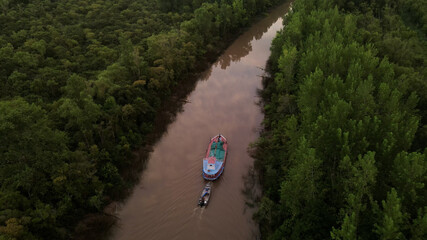 Shot of ship carrying small boat on amazon river surrounded by green rainforest trees during sunset.