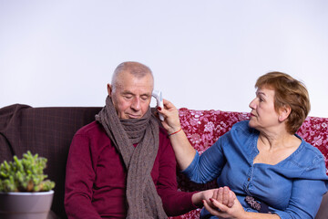 Sad elderly gray senior husband and wife in the apartment, woman measure man fever with non contact thermometer, control heart beat pulse