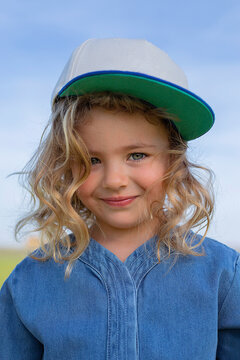 Happy girl in stylish clothes and cap looking at camera while standing on grass on sunny summer day in field