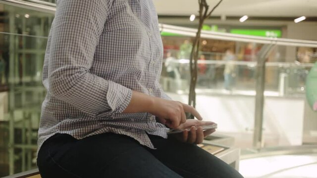 A beautiful young woman sits on a bench in a shopping mall and uses her phone. Internet, communication with loved ones, 4 5g SMS mms, mail. Social communications, rests close up