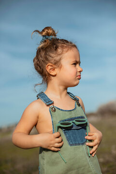 Delighted adorable little girl in overalls standing in meadow and looking away
