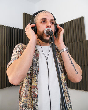 Focused man musician in stylish clothes in headphones standing and singing into microphone while recording song in studio