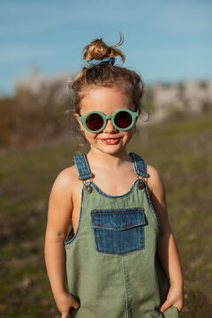 Content little girl in overalls and sunglasses standing in meadow and enjoying summer on sunny day in countryside