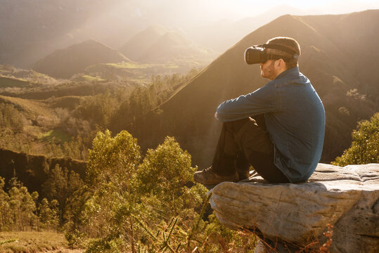 Side view of male traveler in VR glasses interacting with virtual reality while sitting on hill in mountainous terrain at sunset