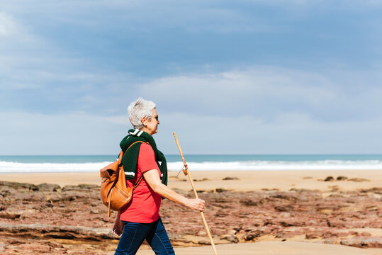 Side view of elderly female backpacker with trekking pole strolling on boulders against stormy ocean under cloudy sky