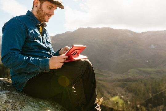 Side view of tranquil male explorer with tablet sitting on rock and enjoying spectacular view of mountains on sunny day