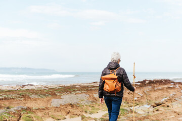 Back view of unrecognizable elderly female backpacker with trekking pole strolling on boulders against stormy ocean under cloudy sky