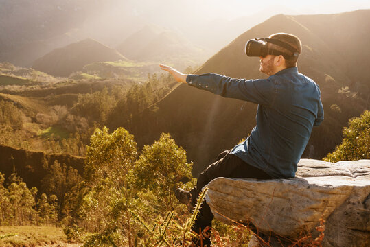 Side view of male traveler in VR glasses interacting with virtual reality while sitting on hill in mountainous terrain at sunset