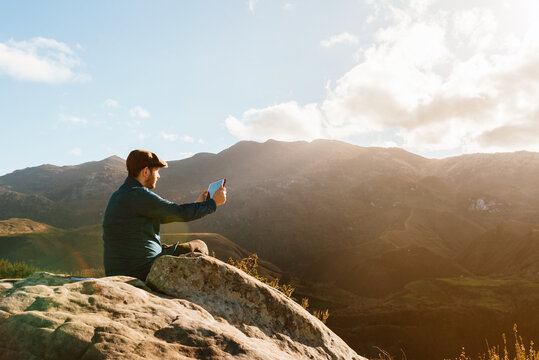 Side view of male explorer sitting on rocky hill and taking picture of mountain range while using tablet