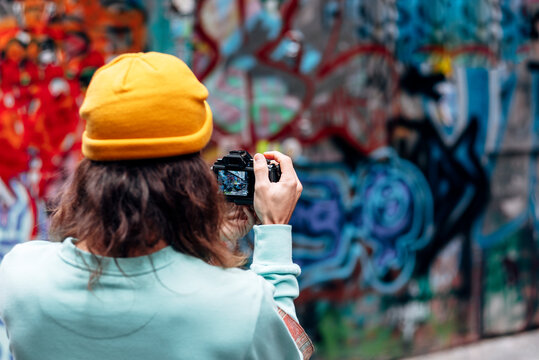 Back view of anonymous hipster male in knitted hat taking photo of graffiti wall on camera in city