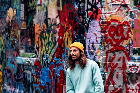 Dreamy hipster young male looking away while standing against graffiti wall in city