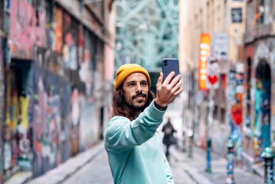 Content male with beard in casual apparel taking self portrait on cellphone in town on blurred background