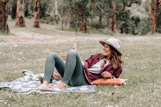 Cheerful female in hat sitting on blanket on meadow in forest and browsing mobile phone while enjoying picnic in Australia