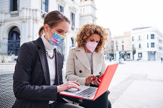 Female entrepreneurs in cloth face masks working on netbook and cellphone on urban bench during COVID 19 pandemic